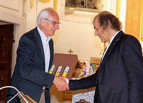 Zwei Männer schütteln sich die Hände und lächeln. Sie halten eine Schachtel mit Flaschen. Der Mann auf der linken Seite trägt eine Brille und einen Anzug. Der Mann auf der rechten Seite trägt einen Anzug und hält eine Schachtel. Hinter ihnen befindet sich eine Holztür und ein Fenster mit einem Kreuz an der Wand.