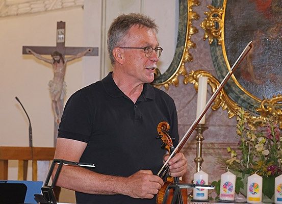 Ein Mann mit Brille spielt Geige in einer Kirche. Er steht vor einem Kreuz und einem Gemälde. Kerzen und Blumen stehen auf dem Tisch vor ihm.