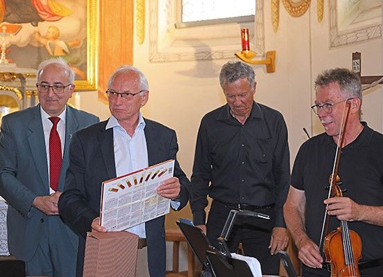 Vier Männer stehen in einer Kirche, einer hält eine Geige, ein anderer liest ein Büchlein. Hinter ihnen hat eine Wand ein Gemälde und ein Kreuz.