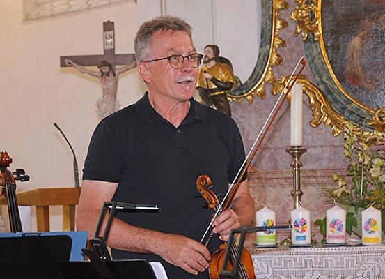 Ein Mann in einem schwarzen Shirt steht in einer Kirche und hält eine Geige. Vor ihm steht ein Mikrofon, und eine Geige ruht auf einem Ständer.