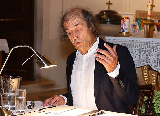 Ein älterer Mann sitzt an einem Tisch, möglicherweise in einer Kirche. Er trägt ein weißes Hemd und eine schwarze Anzugjacke. Eine Brille und eine Lampe stehen auf dem Tisch, mit einem Glas Wasser neben ihm. Hinter ihm steht ein Tisch mit einer weißen Tischdecke und einem Kreuzsymbol.