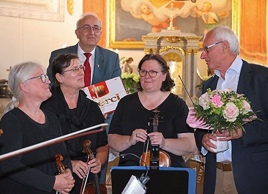 Eine Gruppe von Menschen steht in einem Raum mit Violinen, Notenständern und einem Blumenstrauß. Sie tragen Brillen und formelle Kleidung. Hinter ihnen befindet sich eine Wand mit einem Gemälde.