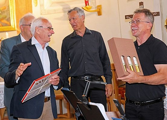 Vier Männer stehen in einem Raum, zwei halten eine Schachtel mit Weinflaschen. Der Mann auf der linken Seite hält ein Buch und spricht, während er gestikuliert.