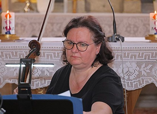 Eine Frau sitzt in einer Kirche und hält einen Notenständer. Sie trägt eine Brille und ein schwarzes Oberteil. Eine Geige ruht auf dem Notenständer. Kerzen stehen auf einem Tisch hinter ihr.