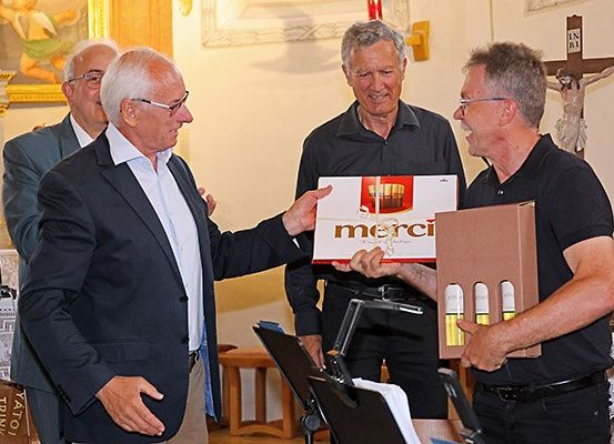 Drei Männer in einer kirchenähnlichen Umgebung, einer hält eine Box mit der Aufschrift Merci, ein anderer Mann zeigt ein Kreuz an der Wand hinter ihm, während der dritte Mann sie ansieht.