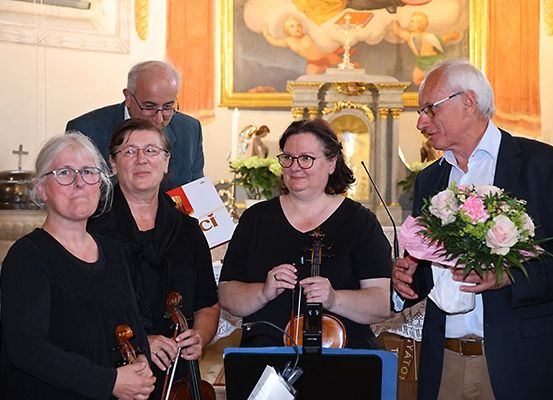 Fünf Personen, darunter zwei Männer, befinden sich in einem Raum mit Musikinstrumenten. Ein Mann hält einen Blumenstrauß. Der Raum hat eine Wand mit einem Gemälde und einer Statue.
