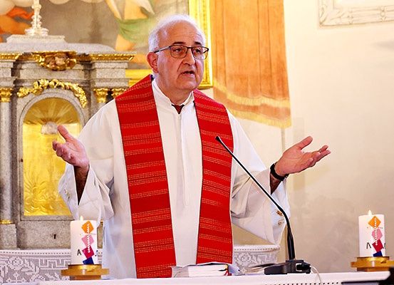 Ein älterer Priester steht an einem Pult, trägt eine rote Stola und eine Brille. Er gestikuliert mit den Händen, während er spricht. Vor ihm steht ein Mikrofon, eine Kerze und ein Buch. Dahinter befindet sich ein goldener Altar mit einem Kreuz.