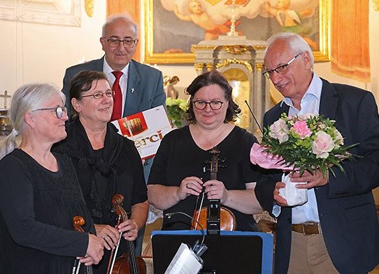 Fünf Personen, darunter zwei Männer und drei Frauen, stehen und halten Violinen. Ein Mann hält einen Blumenstrauß, und vor ihnen steht ein Notenständer. Ein Gemälde schmückt die Wand hinter ihnen.