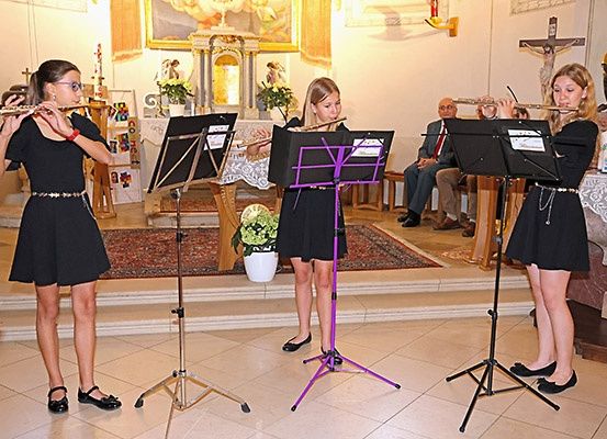 Drei Mädchen spielen Flöten in einer Kirche. Sie stehen auf einem gefliesten Boden mit Notenständern davor. Hinter ihnen sitzt ein Mann auf einer Bank. Ein Kreuz ist an der Wand.