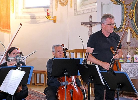 In einem Kirchenumfeld spielen drei Musiker Violine, Flöte und Cello. Sie stehen vor Notenständern und einem Kreuz an der Wand.