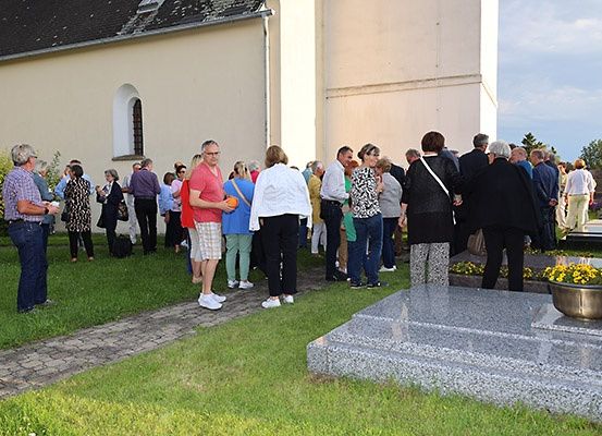 Eine Gruppe von Menschen steht in einer Reihe, möglicherweise wartend, um eine Kirche zu betreten. Sie sind lässig gekleidet, und einige tragen Handtaschen. Ein Grabstein mit gelben Blumen ist in der Nähe.