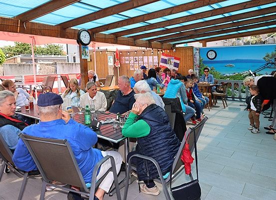 Eine Gruppe von Menschen sitzt an Tischen in einem überdachten Außenbereich mit einer blauen Markise, einige tragen Hüte und Brillen, unter einer Uhr und einer malerischen Kulisse.