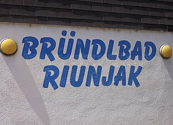 Eine weiße Wand mit blauem Text 'BRUNDLBAD RIUNJAK'. Auf beiden Seiten des Textes befinden sich zwei gelbe Kreise. Die Wand hat eine leichte Schattenbildung auf der linken Seite.