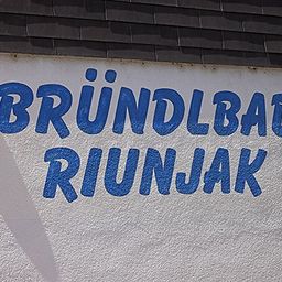 Eine weiße Wand mit blauem Text 'BRUNDLBAD RIUNJAK'. Auf beiden Seiten des Textes befinden sich zwei gelbe Kreise. Die Wand hat eine leichte Schattenbildung auf der linken Seite.