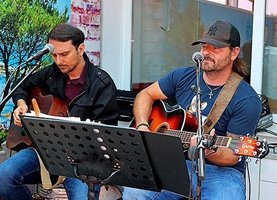 Zwei Männer spielen Gitarre und singen in Mikrofone auf einer Bühne mit einem Hintergrund, der Bäume und Himmel zeigt.