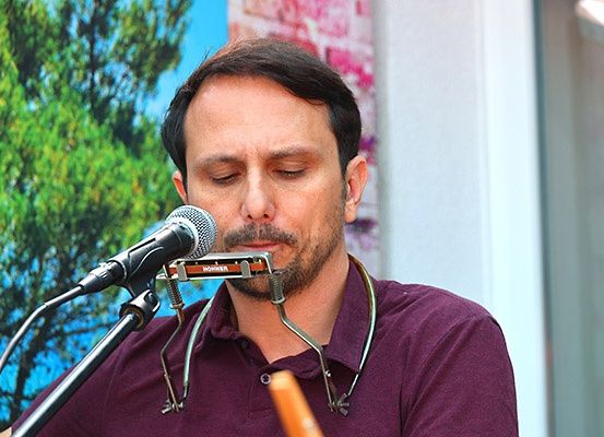 Ein Mann spielt eine Mundharmonika in ein Mikrofon, während er vor einem Mikrofon steht. Er trägt ein weinrotes Hemd und hat Bäume hinter sich.