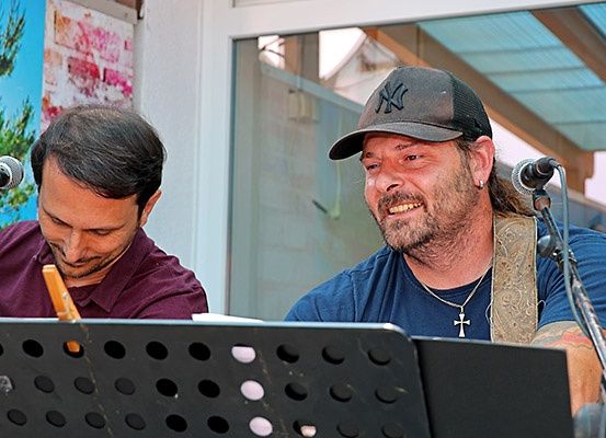 Zwei Männer spielen Musik vor einem Mikrofon. Einer spielt Gitarre und der andere singt. Sie sind draußen.