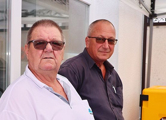 Zwei Männer stehen unter einem Zelt und tragen Sonnenbrillen. Der Mann auf der linken Seite trägt ein weißes Polo-Shirt. Hinter ihnen ist eine Glaswand mit der Reflexion eines Fahrzeugs.