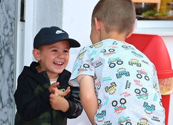 Zwei Jungen, einer in Camouflage und der andere in Monster-Truck-Pyjamas, lächeln und schauen sich in einem Raum an.