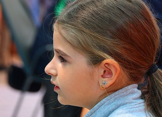 Ein junges Mädchen mit braunen Haaren und roten Highlights blickt nach links, möglicherweise zuhörend. Sie trägt goldene Ohrringe und ein graues T-Shirt. Hinter ihr befinden sich verschwommene Figuren und Stühle.
