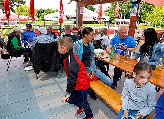 Eine Familie isst in einem Restaurant im Freien. Ein Junge steht, während ein Mädchen auf einer Bank sitzt. Erwachsene unterhalten sich an einem Tisch mit Essen und Getränken.