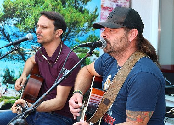 Zwei Männer treten auf der Bühne auf, einer singt in ein Mikrofon, während der andere eine Gitarre spielt. Beide tragen Freizeitkleidung. Hinter ihnen ist eine Wand mit einem Gemälde zu sehen.