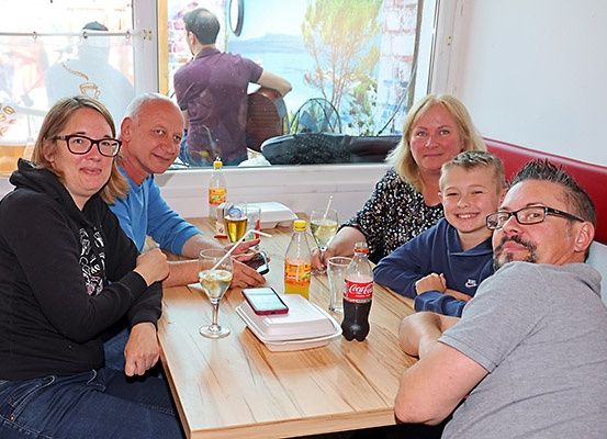 Eine Gruppe von Menschen sitzt lächelnd um einen Tisch in einem Restaurant. Auf dem Tisch stehen Getränke und Essen. Vor dem Fenster spielt ein Mann Gitarre.