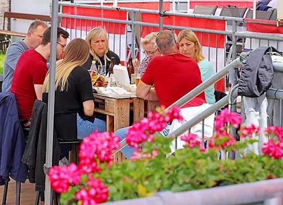 Eine Gruppe von Menschen sitzt um einen Tisch im Freien. Flaschen und Essen stehen auf dem Tisch. Dahinter ist ein Metallzaun und einige Stühle zu sehen.