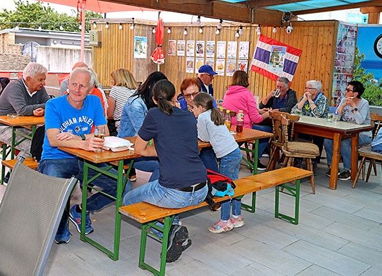 Eine Gruppe von Menschen sitzt an Bänken in einem Café und genießt Getränke und Essen. Die Wände sind mit verschiedenen Postern geschmückt, und eine Fahne hängt in der Nähe. Die Atmosphäre ist locker und entspannt.