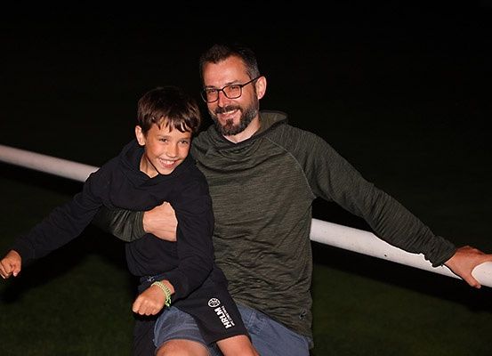 Ein Mann mit Brille und einem grünen Langarmshirt hält einen Jungen in einem schwarzen Sweatshirt und Shorts. Sie stehen auf einem Feld mit einem weißen Pfosten hinter ihnen.