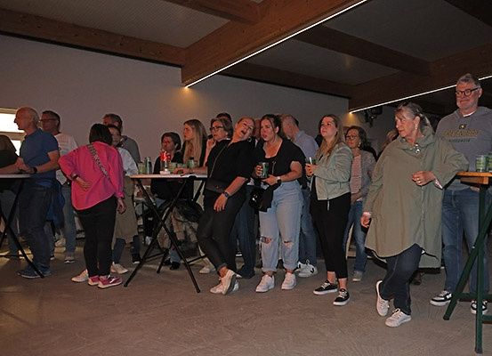 Eine Gruppe von Erwachsenen, sowohl Männer als auch Frauen, versammelt sich in einem Raum, wahrscheinlich bei einer sozialen Veranstaltung. Sie stehen um Tische herum mit Gläsern und Flaschen.