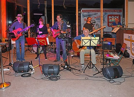 Eine Band spielt bei Nacht auf einer Bühne mit Gitarren, Mikrofonen und Lautsprechern. Der Mann in der Mitte spielt Gitarre sitzend, während andere stehend spielen.