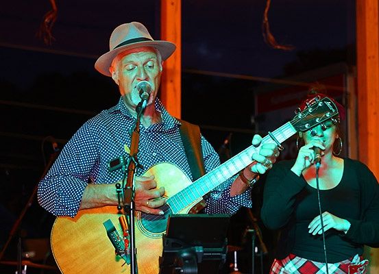 Ein Mann mit Hut spielt Gitarre und singt in ein Mikrofon, während eine Frau in ein anderes Mikrofon singt.