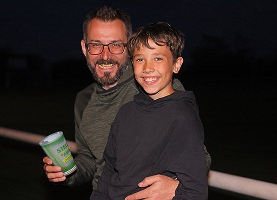 Ein Mann mit Brille und ein Junge in einer Kapuzenjacke stehen draußen bei Nacht. Sie halten einen Becher. Der Junge lächelt. Hinter ihnen steht ein Zaun und ein dunkler Hintergrund.