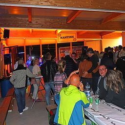 Eine Menschenmenge versammelt sich nachts in einer Cafeteria. Einige stehen, andere sitzen auf Bänken. Es gibt Tische mit Flaschen, Gläsern und Papieren. Ein Schild steht 'KANTIN'. Die Decke hat Holzbalken und Lichter.