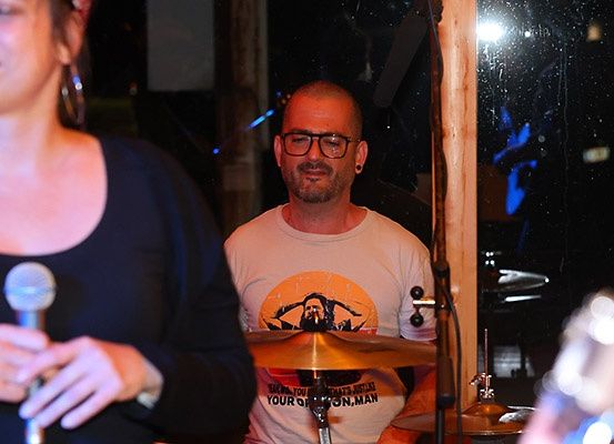 Ein Mann spielt die Trommeln, während eine Frau in ein Mikrofon singt in einem schwach beleuchteten Raum.