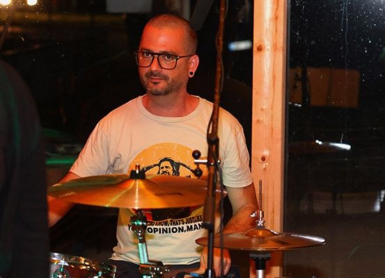 Ein Mann spielt die Trommeln in einem Raum, trägt eine Brille und ein weißes T-Shirt mit einem grafischen Design.