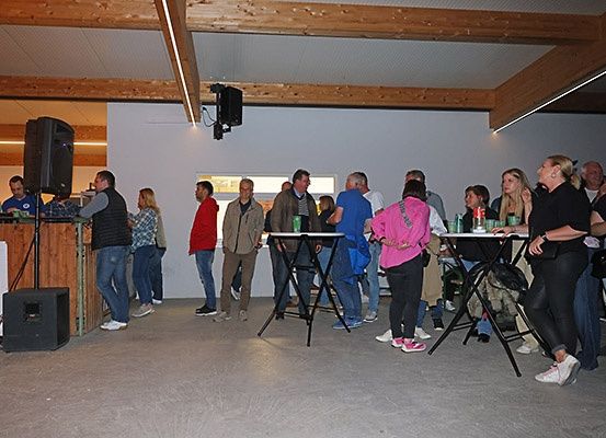 Eine Gruppe von Menschen versammelt sich in einem Raum mit Tischen und Stühlen. Eine Person steht hinter einer Theke mit einem Lautsprecher. Der Raum hat Holzbalken und eine weiße Wand.