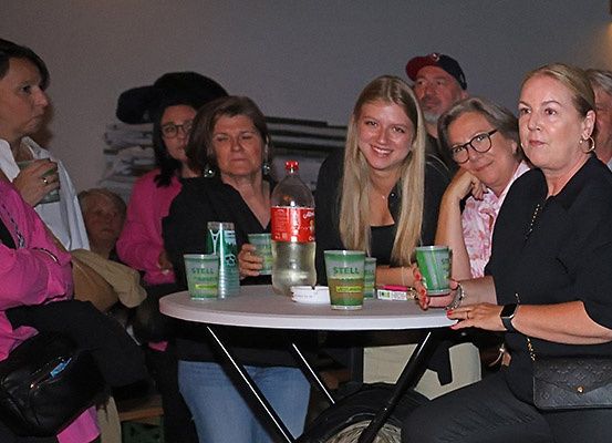 Mehrere Personen stehen um einen Tisch mit Getränken und Snacks. Eine Frau in einem rosa Shirt hält ein Getränk. Eine andere Frau lächelt in die Kamera.