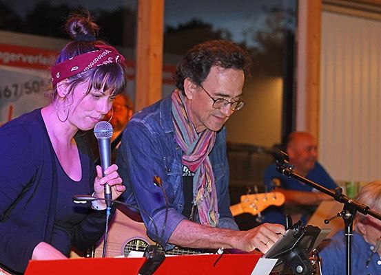 Ein Mann und eine Frau treten mit Musikinstrumenten auf der Bühne auf. Die Frau singt in ein Mikrofon, und der Mann spielt Gitarre. Hinter ihnen sitzt eine Person und spielt Flöte. Ein Banner hängt an der Wand.