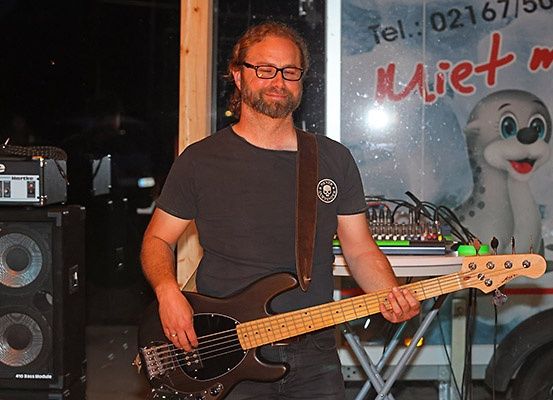 Ein Mann mit Brille und schwarzem T-Shirt spielt lächelnd eine Bassgitarre vor einer Wand mit einem Comic-Seehund und einer Telefonnummer.