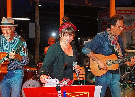 Eine Band spielt bei Nacht. Der Mann links spielt Gitarre und trägt einen Hut. Die Frau in der Mitte singt in ein Mikrofon. Der Mann rechts spielt Gitarre. Dahinter steht ein Schlagzeug und ein Plakat.