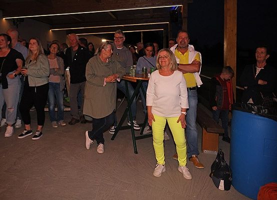 Eine Gruppe von Menschen ist in einem schwach beleuchteten Bereich versammelt, wahrscheinlich für eine Veranstaltung. Einige stehen, andere sitzen, und zwei Frauen tanzen. In der Mitte steht ein Tisch mit Gläsern. Ein Junge und ein erwachsener Mann stehen in der Nähe eines Mülleimers.