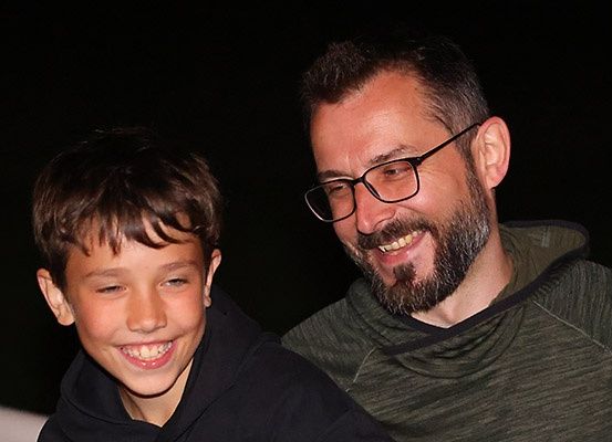 Ein lächelnder Junge und ein Mann mit Brille posieren nachts für ein Foto. Der Junge trägt eine schwarze Kapuzenjacke, und der Mann trägt eine graue Kapuzenjacke. Sie haben beide fröhliche Ausdrücke.
