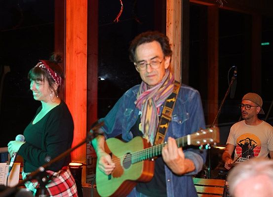Ein Mann mit Brille und Schal spielt Gitarre, während eine Frau ein Mikrofon hinter ihm hält. Ein anderer Mann spielt Schlagzeug im Hintergrund.