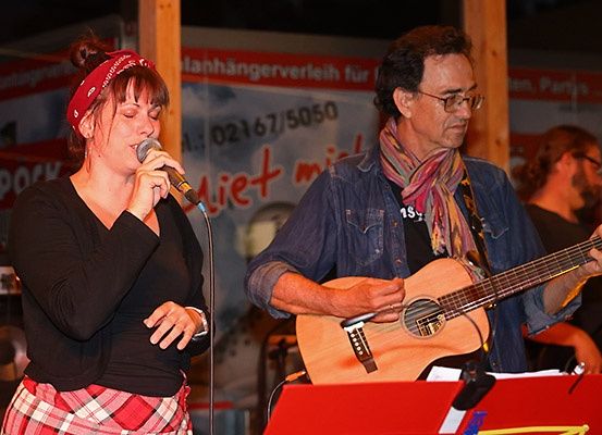 Eine Frau mit einem roten Stirnband singt in ein Mikrofon, während ein Mann Gitarre spielt. Sie sind auf einer Bühne mit einem Banner im Hintergrund.