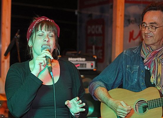 Eine Frau singt in ein Mikrofon auf der Bühne bei Nacht, neben ihr ein Gitarrist, ein Banner im Hintergrund und ein Schlagzeug hinter ihnen.