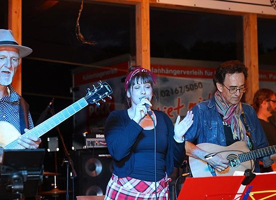 Eine Band spielt auf der Bühne, eine Frau singt in ein Mikrofon, zwei Männer spielen Gitarre und verschiedene Instrumente im Hintergrund.