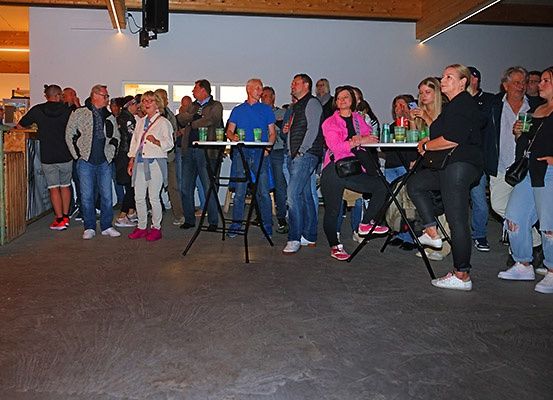 Eine Gruppe von Menschen steht in einem Raum mit Tischen und Stühlen. Einige halten Getränke und zwei Frauen sitzen. Der Raum hat weiße Wände und eine Holzdecke.
