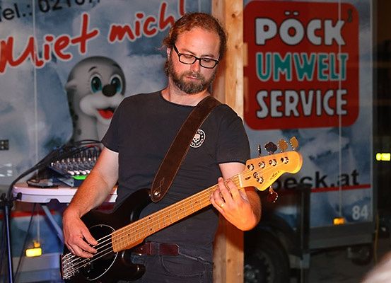 Ein Mann mit Bart und Brille spielt eine Bassgitarre vor einem Banner mit einem Cartoon-Seehund und rotem Text.